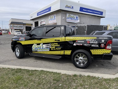 Black work truck with yellow tire tread graphics across the side and logos for tire and wheel brands. It is a tire and auto service truck and is parked in front of Clark's Tire & Automotive in Spokane, WA